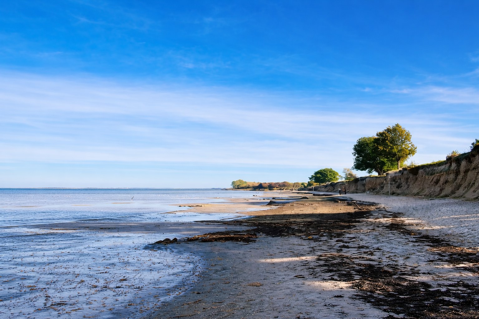 Weiter Sandstrand bei Ebbe mit ruhigem Meer, blauem Himmel und bewachsener Steilküste im Hintergrund