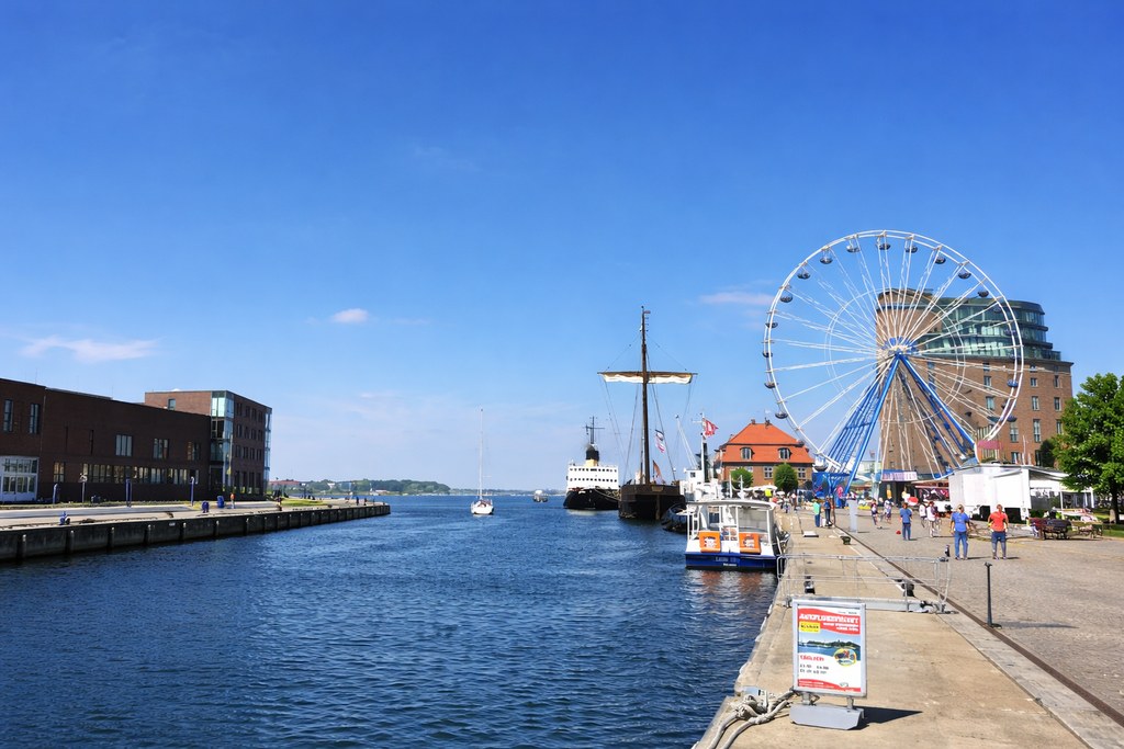 Blick auf eine lebendige Hafenpromenade mit Wasser, Schiffen und großem Riesenrad bei blauem Himmel an einem sonnigen Tag