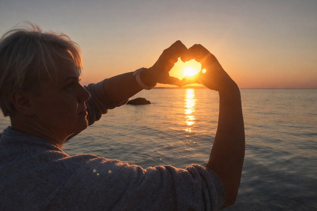 Person formt mit den Händen ein Herz um die untergehende Sonne am Meer bei warmem Abendlicht